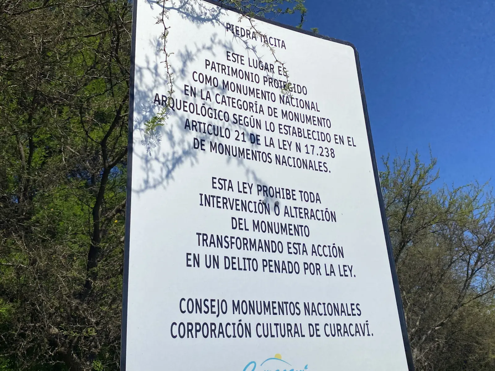 Cartel oficial blanco de la Piedra Tacita que avisa que el lugar es patrimonio protegido como Monumento Nacional, con el nombre del Consejo de Monumentos Nacionales y de la Corporación Cultural de Curacaví