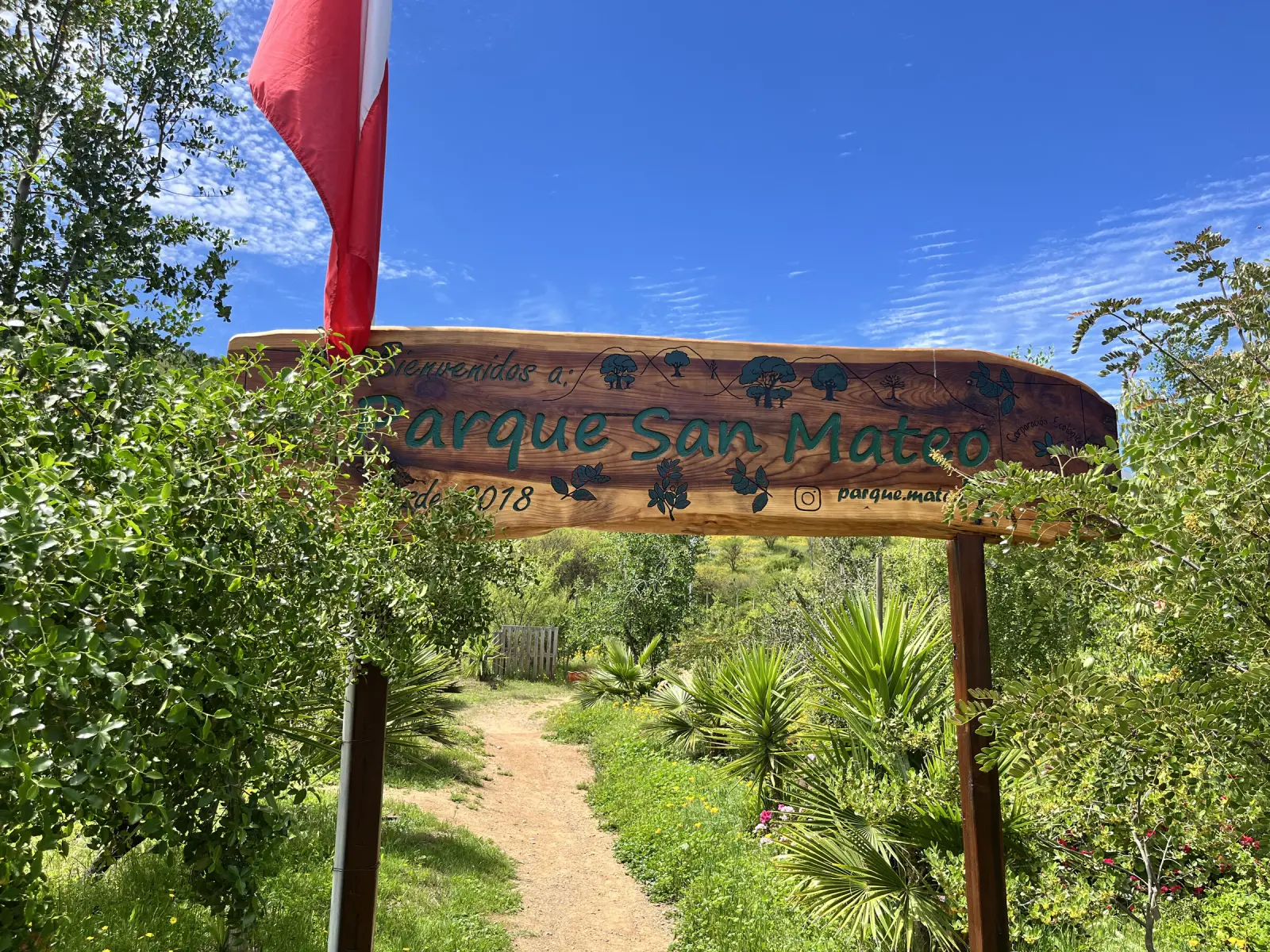 Portal de madera con el letrero «Bienvenidos a Parque San Mateo» y la bandera de Chile, sobre el sendero de entrada al parque