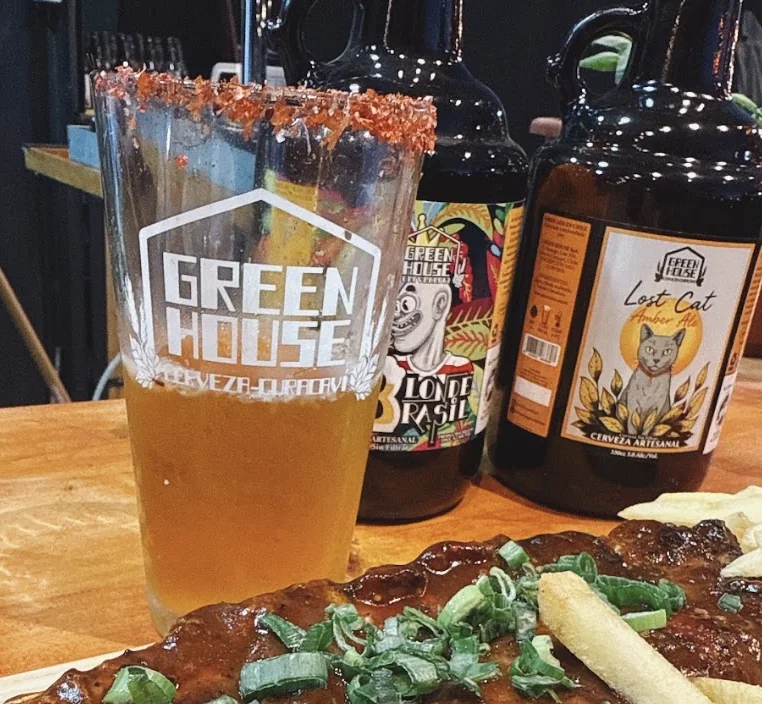 Vaso con el logo de Green House junto a botellas de cerveza artesanal y un plato de comida